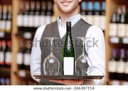 Marvelous taste experience. Close-up of a tray with a bottle of wine and two glasses on it held by a smiling sommelier in blurry