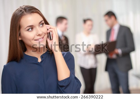 Business calls. Confident beautiful female manager talking over the phone with a smile while her colleagues standing in blurry on the background