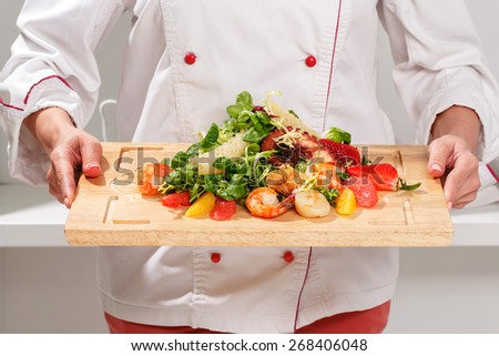 Close-up of a cook holding a cutting board with salad with octopus, shrimps, scallops, strawberries, orange, mash-salad, frisee