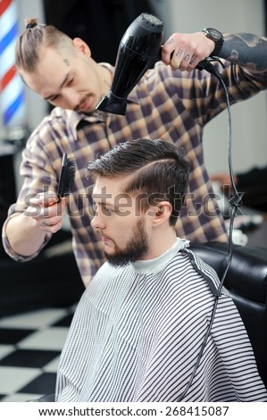 Making a hairdo. Hairstylist drying with a hairdryer and a comb male client sitting in chair at barbershop