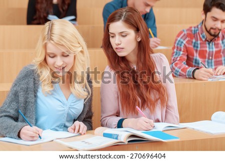 Cheating on test. Young beautiful red haired student peeping to the exercise book of her friend sitting by her side at class