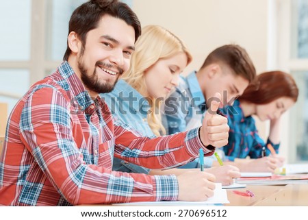Best time studying. Young smiling male student showing his thumb up sitting in class with his peers