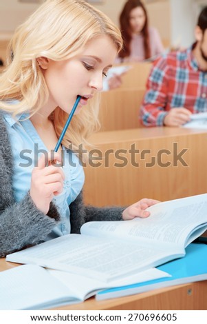 Study hard. Vertical shot of a young concentrated female student biting a pen while looking at her exercise book sitting in class