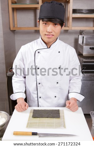 Master class in sushi cooking. Young Japanese chef looking at a camera standing before the table with a knife, bamboo mat and seaweed sheet on it