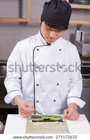 Skillful cook. Young smiling Japanese cook in white uniform preparing sushi on a bamboo mat