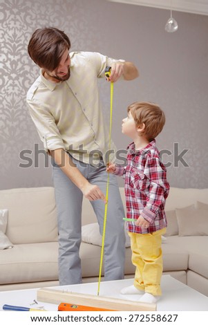 Growing big. Father tracks with a ruler height of his son stranding on a small table in a living room