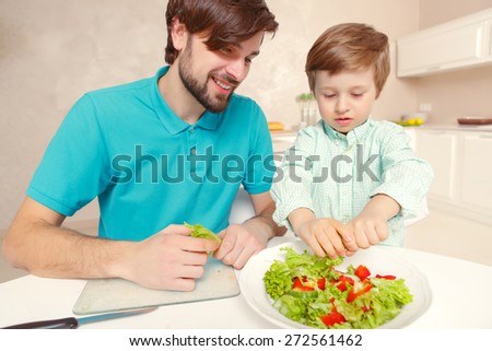 Family time. Young smiling father and his son cooking fresh vegetable salad at home kitchen