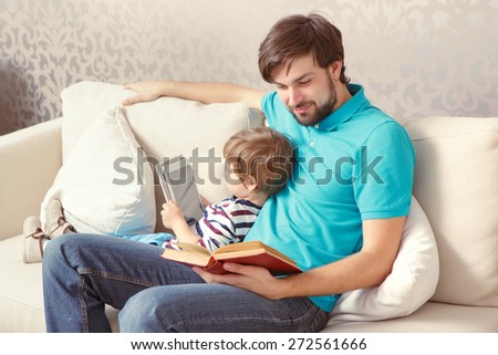Different generations. Young father reading a book while his lovely son using a tablet device sitting at the home couch