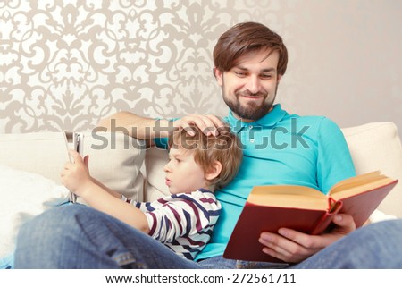 Different generations. Young father reading a book while his lovely son using a tablet device sitting at the home couch