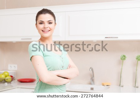 Serious attitude. Young smiling lady standing in kitchen with her arms folded across her chest