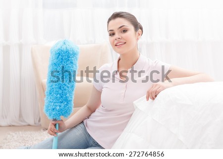 Feel free. Young lady sitting on white sofa with blue cleaning sweep on background of white curtain