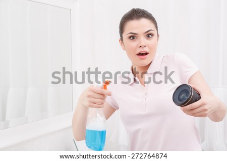 Why not. Young hilarious girt with amazed look trying to clean object-glass on white background in front of mirror