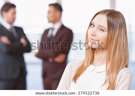 Office workers. Blond young business woman standing in foreground giving a wink, her co-workers discussing business matters in the background