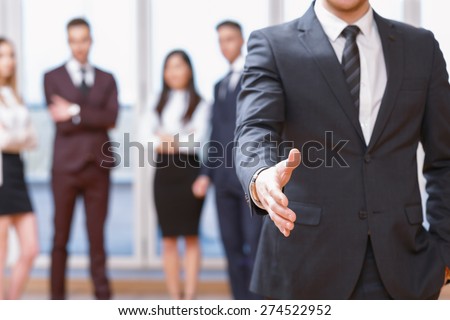 Nice to meet you.  Young business man standing in foreground extending hand for a handshake, his co-workers discussing business matters in the background
