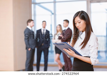 Office workers.  Asian young business woman standing in foreground looking at the tablet seriously, her co-workers discussing business matters in the background