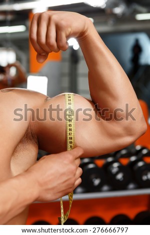Proud of results.  Really muscular man measuring size of his biceps with help of soft tape measure in gym.