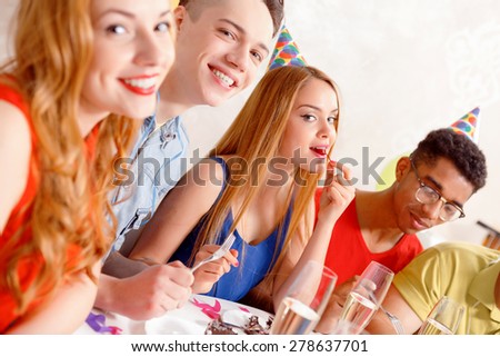 Birthday party. Young happy people wearing cone caps sitting in a row at the table while beautiful blond girl biting a cherry selective focus