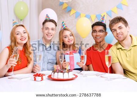 Birthday party. Young happy people wearing cone caps sitting at the table holding glass of champagne, present while the cake standing on the table in a decorated with balloons room