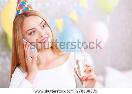 Birthday call. Young beautiful blond girl wearing cone cap taking congratulations on her birthday talking on her mobile phone and holding glass of champagne