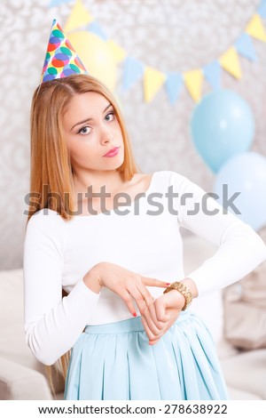 Friends are late. Young beautiful blond girl wearing cone cap pointing at her watches with a sad face because her guests are late for the birthday party