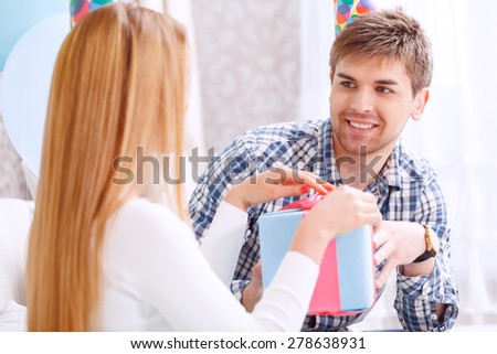 Portrait of a young man presenting a birthday gift in a blue box tied with red ribbon to his girl friend smiling celebrating in a decorated room