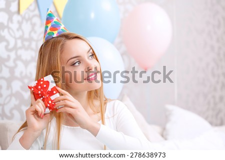 Portrait of a young beautiful blond girl wearing cone cap sitting on a couch and trying to hear what is inside her birthday present