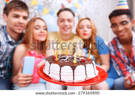 Celebration. Young happy boy holding his birthday cake while his friend smiling and holding presents in their hands and looking at the cake selective focus