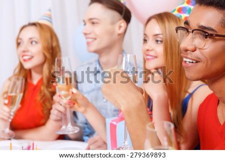 Birthday party. Young happy people wearing cone caps sitting in a row at the table holding glass of champagne, present while the cake standing on the table in a decorated with balloons room