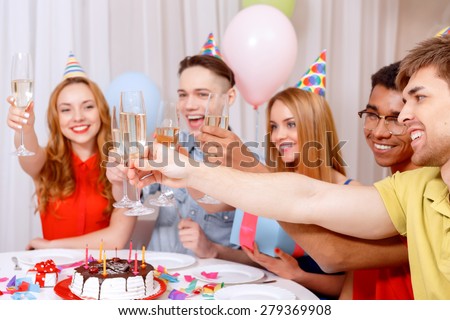 Birthday party. Young happy people wearing cone caps sitting at the table laughing holding and clinking glasses of champagne near the birthday cake