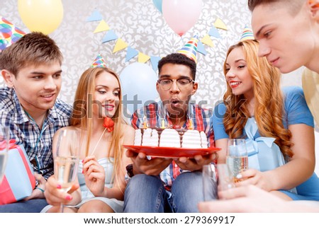 Celebration. Young happy boy blowing candles from his birthday cake making a wish while his friends smiling and holding glasses of champagne in their hands and looking at the cake