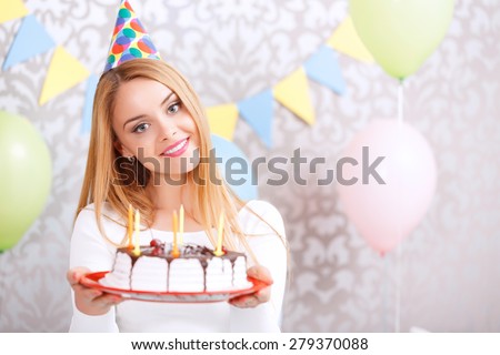 Portrait of a young beautiful blond girl wearing cone cap holding a red plate with birthday cake with candles in the light decorated room