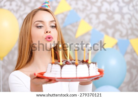 Portrait of a young beautiful blond girl wearing cone cap holding a red plate with birthday cake and blowing candles making a wish