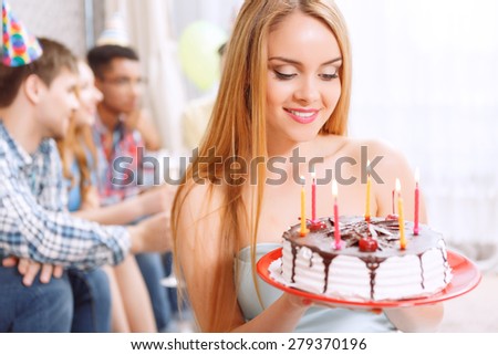 Sweetness. Portrait of a young beautiful blond girl wearing cone cap looking at her birthday cake with burning candles on it and her friends on the background celebrating