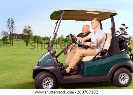 Moving forward. Side-draw of smiling old and young golf player driving cart on course.