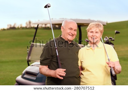 Satisfied with game. Two lovely senior people standing with golf clubs on background of cart on course.
