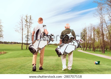 Father and son. Back view of old and young golf players walking on course with golf equipment.