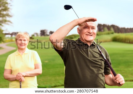 Into distance. Senior man standing on course near his wife holding his hand near his forehead and looking into the distance