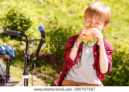 So hungry. Funny little boy sitting on bench near his bicycle in park and biting off piece of sandwich