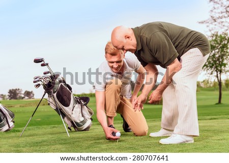 Preparatory work. Young and old goler  putting ball on tee on course on background of bag.