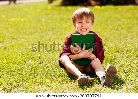 It is mine. Little smiling boy sitting on grass and gently hugging big green book.