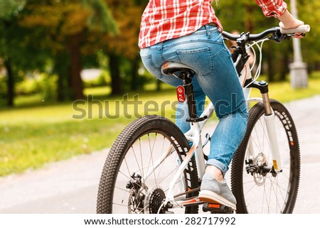 Photo of a girl riding a sport bicycle wearing a red checkered shirt jeans and grey sneakers in a green park, back view