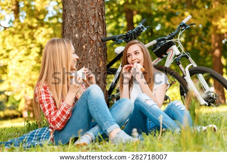 Two young beautiful girls with long hair sitting on a blue checkered mat under a tree eating sandwiches and smiling, while their bikes standing near in a green park