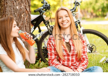 What a wonderful picnic. Two young beautiful girls with long hair sitting on a blue checkered mat under a tree smiling and drinking fruit juice, their bikes standing near in a green park