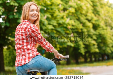 :  Portrait of a young beautiful blond girl with long hair posing sitting on her mountain bicycle smiling wearing a red checkered shirt, in a green park