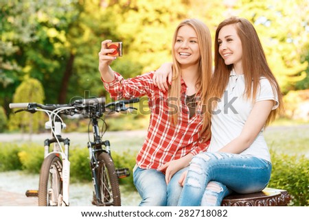 Two young beautiful happy girls sitting on a bench in a green park smiling and making selfie on a colorful smartphone, their sport bikes standing near, selective focus
