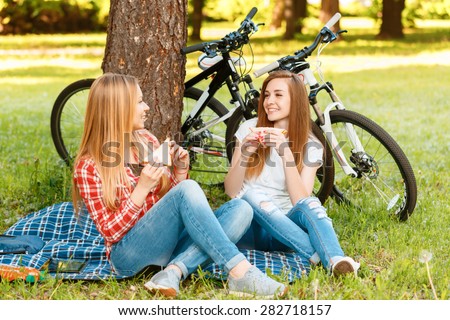 Two young beautiful girls with long hair sitting on a blue checkered mat under a tree eating sandwiches and smiling, while their bikes standing near in a green park