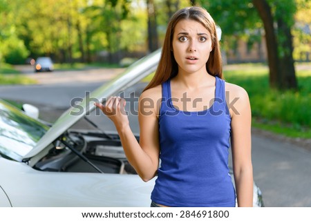 Feeling confusion. Portrait of embarrassed young woman pointing on broken car behind her.