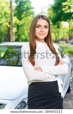 Self-confidence. Pleasant youthful girl standing next to new white car with crossed arms.