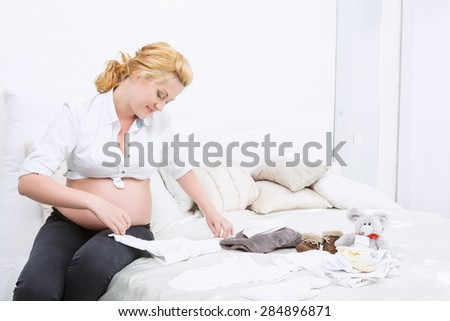 Nice worries. Pregnant young women  carefully folding clothes while sitting on bed