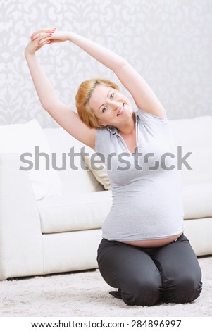 Enjoying sport. Pregnant young  woman doing side stretching exercise and smiling while sitting on the carpet.